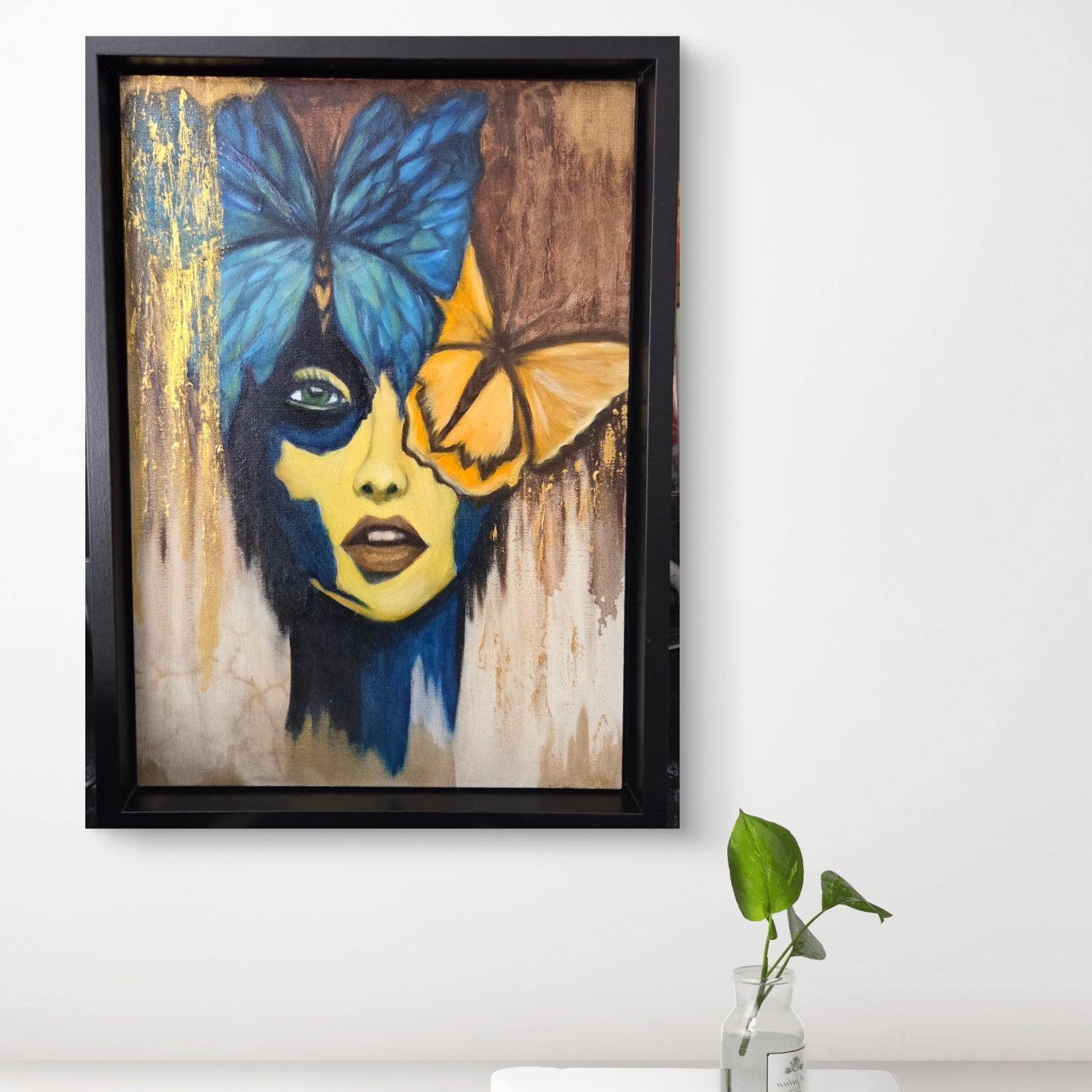 Framed artwork of a woman's face with butterflies on a white wall 