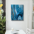 Abstract blue painting with gold accents on a white wall above a desk with a laptop and a cup.